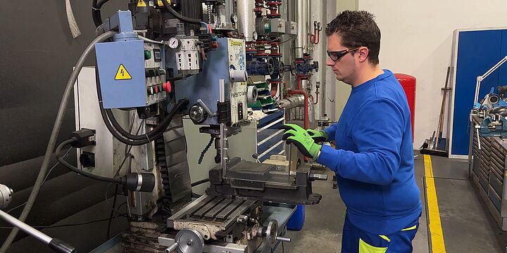 Ein Mann mit blauem Pullover, Schutzbrille und Arbeitshandschuhen führt an einer Fräsmaschine einen Sicherheits-Check durch