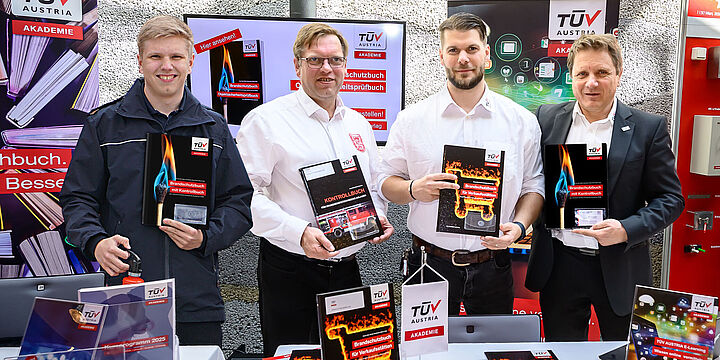 ©TÜV AUSTRIA Akademie 4 Männer stehen hinter einem Ausstellertisch und halten das neue Brandschutzbuch in die Kamera