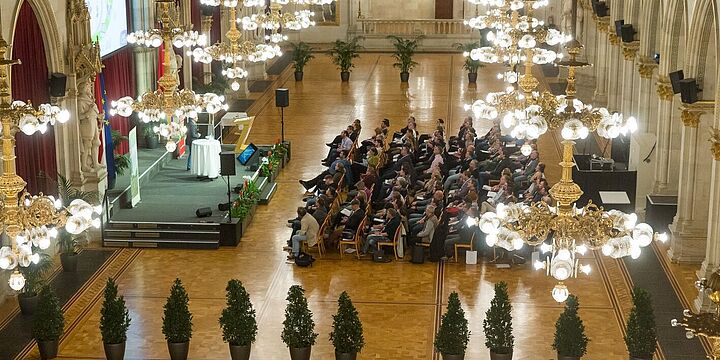 Festsaal im Wiener Rathaus. Publikum sitzt vor Bühne und hört sich Vortrag an.